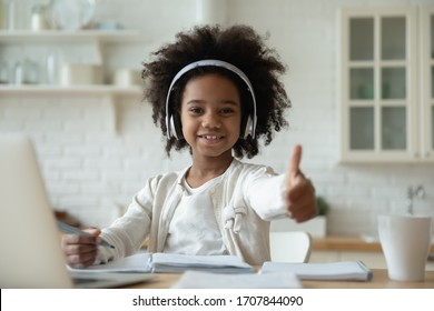 Portrait of happy little African American girl pupil in headphones study online using laptop at home, smiling small biracial child show thumb up recommend class or lesson, homeschooling concept - Powered by Shutterstock