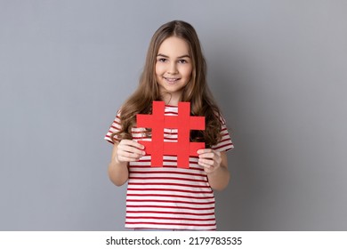 Portrait of happy little girl wearing striped T-shirt holding hashtag symbol, promoting viral topic in social network, tagging blog trends. Indoor studio shot isolated on gray background.