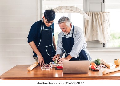 Portrait Of Happy Love Asian Family Senior Mature Father And Young Adult Son Having Fun Cooking Together And Looking For Recipe On Internet With Laptop Computer To Prepare The Yummy Eating Lunch