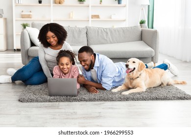 Portrait of happy loving black family lying on floor carpet with labrador, using laptop and watching video or movie, browsing internet, spending time together at home in living room