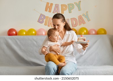Portrait of of happy loving woman sitting on sofa with her infant daughter and holding cake with candles, kissing cute baby, expressing positive emotions, celebrating first baby birthday.