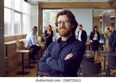 Portrait Of Happy Male Business Teacher And Professional Coach. Handsome Long Haired Bearded Man In Blue Shirt And Eyeglasses Standing In Office After Corporate Training Class For Team Of Employees