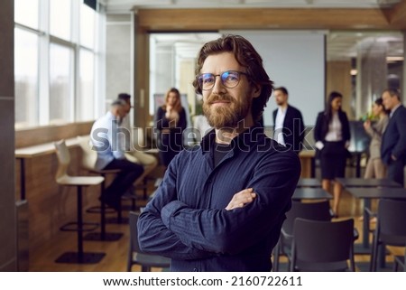 Portrait of happy male business teacher and professional coach. Handsome long haired bearded man in blue shirt and eyeglasses standing in office after corporate training class for team of employees