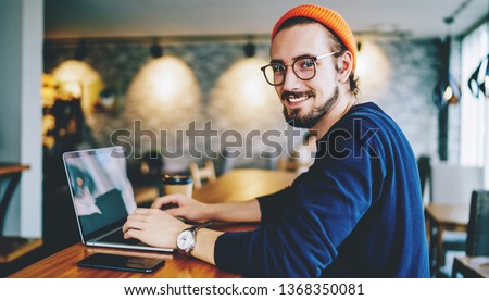 Portrait of happy male freelancer in optical eyewear smiling at camera during break from working online, positive cheerful hipster blogger sitting on publicity area with modern laptop computer