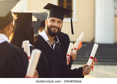 Portrait of happy male graduate holding his diploma standing in row with his groupmates near university. Handsome bearded student smiling and looking at camera during college graduation