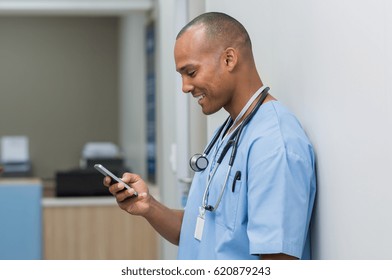 Portrait of happy male nurse reading message at mobile phone. Young african surgeon text messaging through cell phone. Man using smart phone in hallway at hospital. - Powered by Shutterstock