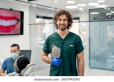 Portrait of a happy male professional dentist with digital tablet standing in his dental office and smiling at camera. Nurse and patient in dentist's chair in background. Toothy smile on a monitor. - Powered by Shutterstock