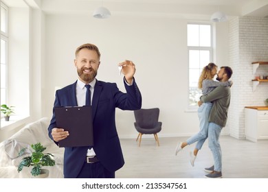 Portrait Of Happy Male Real Estate Agent Show Keys To Home, Excited Couple Celebrate Relocation Moving On Background. Smiling Relator Congratulate Clients With House Buy. Rental, Realty Concept.