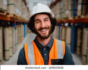 Portrait Of Happy Male Worker In Warehouse Wearing Orange Vest And White Helmet Standing Between Shelves