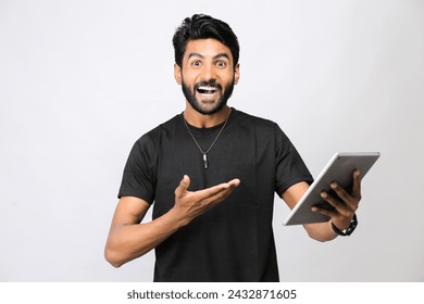 a portrait of a happy man with a beard and a fashionable hairstyle, dressed in casual clothes, stands with a tablet and shows his hand to the pointing to tablet, Isolated on a plane background - Powered by Shutterstock