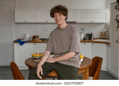 Portrait of a happy man with brown curly hair wearing casual clothing  looking at the camera and smiling while sitting on a table in modern white kitchen and enjoying life indoors. Lifestyle concept. - Powered by Shutterstock