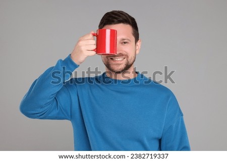 Portrait of happy man covering eye with red mug on grey background