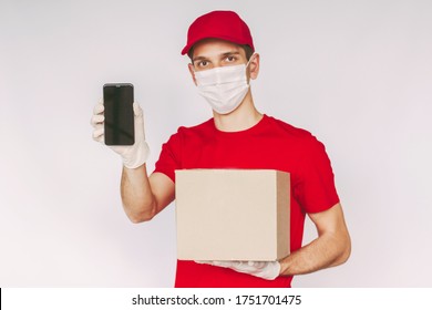 Portrait Happy Man Delivery Service Employee In Medical Face Mask, Protective Gloves Hold Mobile Phone, Parcel Box With Empty Blank Copy Space. Young Delivery Guy Carry Cardboard Box And Smart Phone