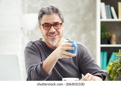 Portrait Of Happy Man At Home Sitting At Desk, Working, Looking At Camera. Happy Smile, Grey Hair, Beard, Glasses. Portrait Of Mature Age, Middle Age, Mid Adult Man In 50s.