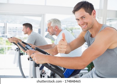 Portrait Of Happy Man On Exercise Bike Gesturing Thumbs Up At Gym