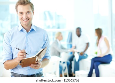 Portrait Of Happy Man With Pen And Notepad Looking At Camera In Working Environment