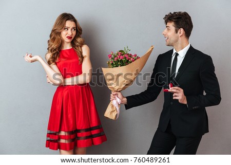 Portrait of a happy man proposing to a unsatisfied girl with flowers and an engagement ring over gray wall background