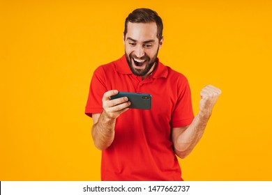 Portrait of happy man in red t-shirt smiling and holding smartphone while playing video games isolated over yellow background - Shutterstock ID 1477662737