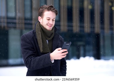 Portrait Of Happy Man, Young Guy Businessman In Formal Clothes Is Looking At Screen Of His Cell Mobile Phone, Using Smartphone And Smile Outdoors At Winter Snowy Snow Cold Day