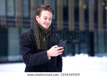 Portrait of happy man, young guy businessman in formal clothes is looking at screen of his cell mobile phone, using smartphone and smile outdoors at winter snowy snow cold day