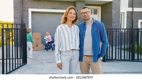 Portrait of happy married couple with children standing in hugs at new house and smiling cheerfully. Kids on background.