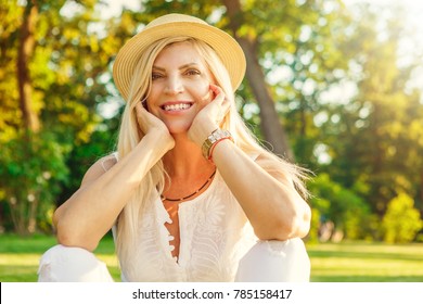 Portrait Of A Happy Mature Beautiful Woman In A Hat Smiling Cupping Her Face Sitting On The Grass At The Park Enjoying Warm Summer Day Outdoors Copyspace Happiness Vitality Positivity Wellbeing