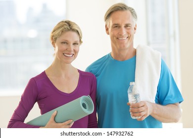 Portrait Of Happy Mature Couple With Exercise Mat, Water Bottle And Towel In Health Club