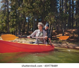 Portrait Of Happy Mature Couple Having Fun At The Lake. Woman Paddling Kayak With Man Pushing From Behind On A Summer Day.