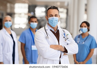 Portrait Of Happy Mature Doctor Standing In Corridor With Medical Team At Hospital Wearing Surgical Face Mask Due To Covid. Smiling General Practitioner With Crossed Arms Looking At Camera.