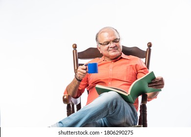 Portrait of a happy mature Indian or asian man reading a book while sitting on rocking chair sipping tea or coffee from cup - Powered by Shutterstock