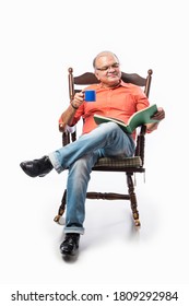 Portrait of a happy mature Indian or asian man reading a book while sitting on rocking chair sipping tea or coffee from cup