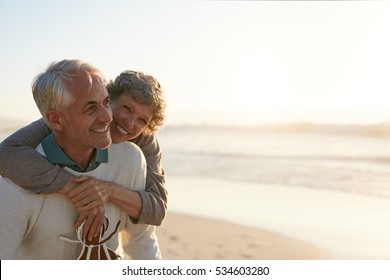 Portrait Of Happy Mature Man Being Embraced By His Wife At The Beach. Senior Couple Having Fun At The Sea Shore.