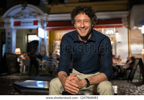 Portrait of happy\
mature man in formal clothing sitting on bench and looking at\
camera at night. Senior business man in smart casual sitting\
outside restaurant during night\
time.