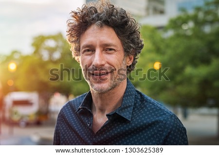 Portrait of happy mature man looking at camera outdoor. Senior businessman with beard feeling confident at sunset. Closeup face of business man smiling with city in background.