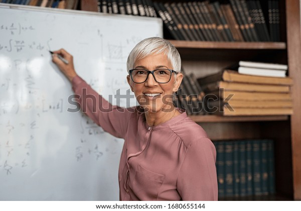 Portrait of happy mature professor teaching\
mathematics to students in a library. Senior smiling woman solving\
math problem while writing on white board. Portrait of tutor\
looking at camera.