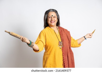 Portrait Of Happy Mature Senior Indian Woman Holding Rolling Pin And Wooden Spoon Isolated On White Background. Home Maker, Kitchen Concept.