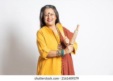 Portrait Of Happy Mature Senior Indian Woman Holding Rolling Pin And Wooden Spoon Isolated On White Background. Home Maker, Kitchen Concept.