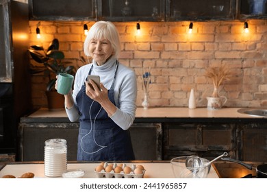 Portrait of happy mature woman chilling in kitchen while cooking, copy space. Smiling senior woman in apron with cup of tea coffee listening music on mobile phone in kitchen - Powered by Shutterstock
