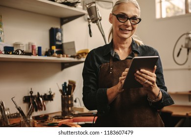 Portrait of happy mature woman jewelry maker using digital tablet in her workshop. Smiling senior woman in goldsmith workshop with tablet pc. - Powered by Shutterstock