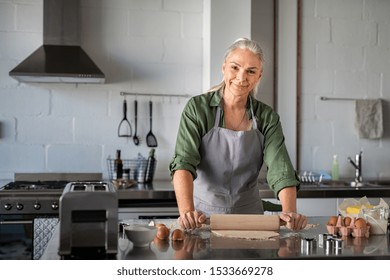 Portrait of happy mature woman making dough at home. Portrait of housewife flattening cookie dough using wooden rolling pin in kitchen while smiling and looking at camera. Old woman preparing cookies. - Powered by Shutterstock