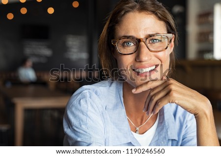 Portrait of happy mature woman wearing eyeglasses and looking at camera. Closeup face of smiling woman sitting in cafeteria with hand on chin. Successful lady in a cafe pub.
