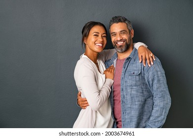 Portrait of happy mid adult couple embracing and looking at camera standing against gray background. Mature indian man in love standing on grey wall while hugging his hispanic woman with copy space.