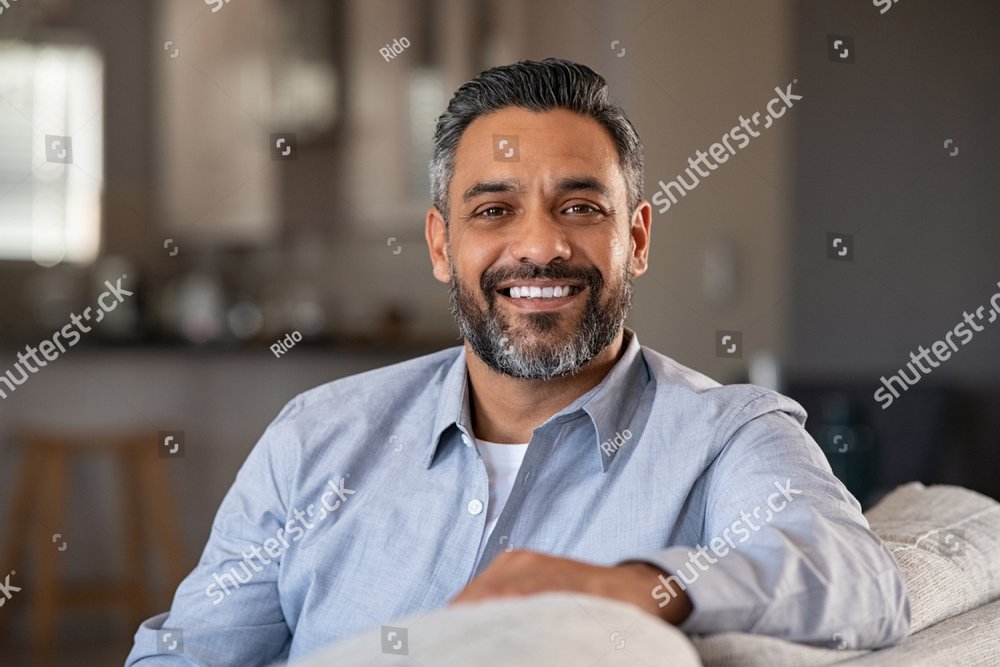 Portrait of happy mid adult man sitting on sofa at home. Handsome latin man in casual relaxing on couch and smiling. Cheerful indian guy looking at camera. 