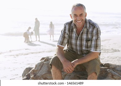 Portrait Of Happy Middle Aged Man Sitting On Rock While Family Enjoying In Background At Beach