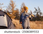 Portrait of happy middle aged woman holding coffee cup while standing near tent. 40s caucasian woman having tea at campsite on a sunny day