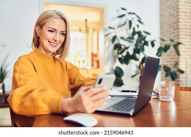 Portrait of happy middle aged  woman using phone and laptop sitting at table working from home - Powered by Shutterstock