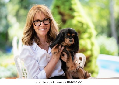 Portrait Of Happy Middle Aged Woman Holding Her Cute Puppy And Sitting In A Lounge Chair While Relaxing At The Backyard At Home. 