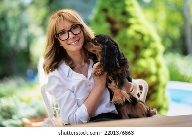 Portrait Of Happy Middle Aged Woman Holding Her Cute Puppy And Sitting In A Lounge Chair While Relaxing At The Backyard At Home. 