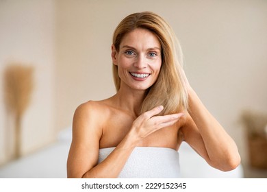 Portrait of happy middle aged woman touching her soft hair, standing in bedroom and smiling at camera. Lady enjoying beauty routine, caring for herself