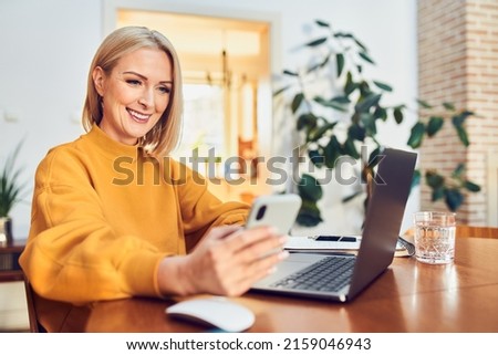 Portrait of happy middle aged  woman using phone and laptop sitting at table working from home